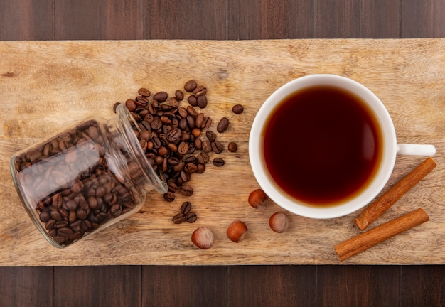 Top view of coffee beans spilling out of glass jar and cup of tea with cinnamon and nuts on cutting board on wooden background