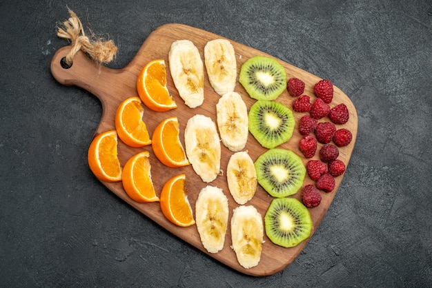 Free photo top view of collection of chopped fresh fruits on a wooden cutting board on black table in vertical view