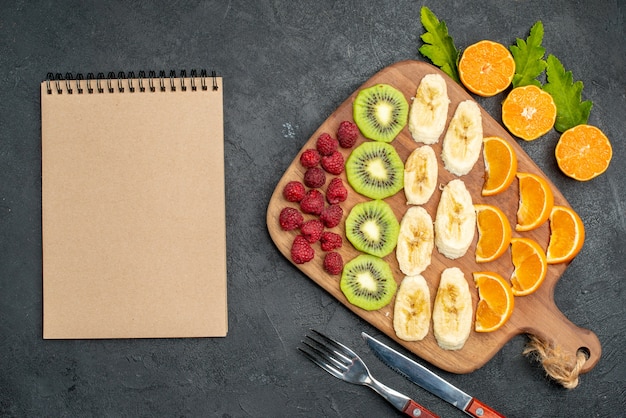 Top view of collection of chopped fresh fruits on a wooden cutting board and spiral notebook on black table