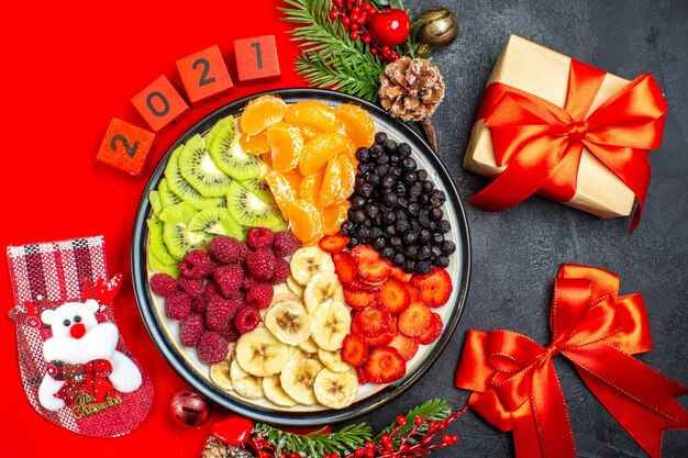 Top view of collection of fresh fruits on dinner plate decoration accessories fir branches and numbers on a red napkin and red ribbon and gift