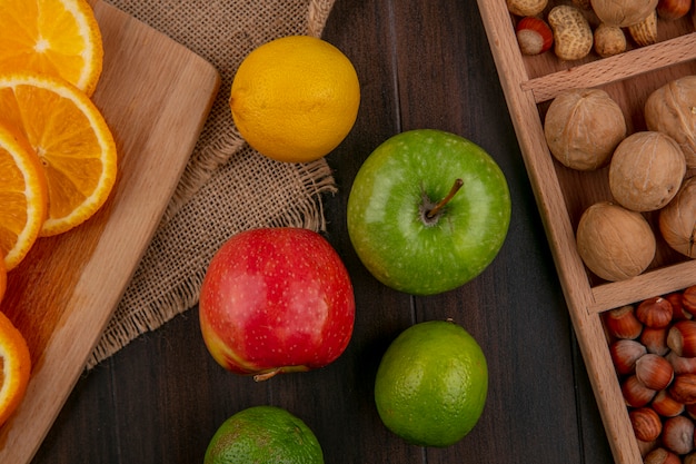 Free photo top view of colored apples with sliced orange on a board with lemon blackcurrant and nuts on a wooden surface