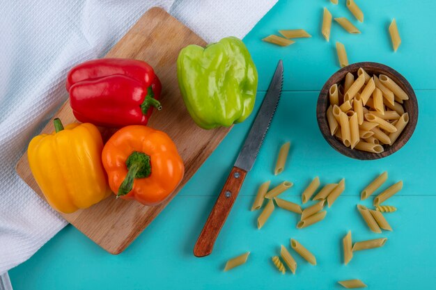 Top view of colored bell peppers on a cutting board with raw pasta and a knife on a turquoise surface