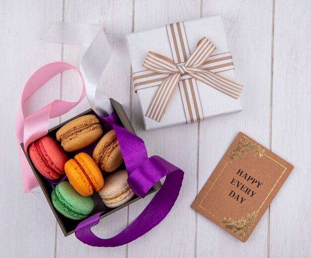 Top view of colored macarons in a box with colored bows and a gift box with a book on a white surface