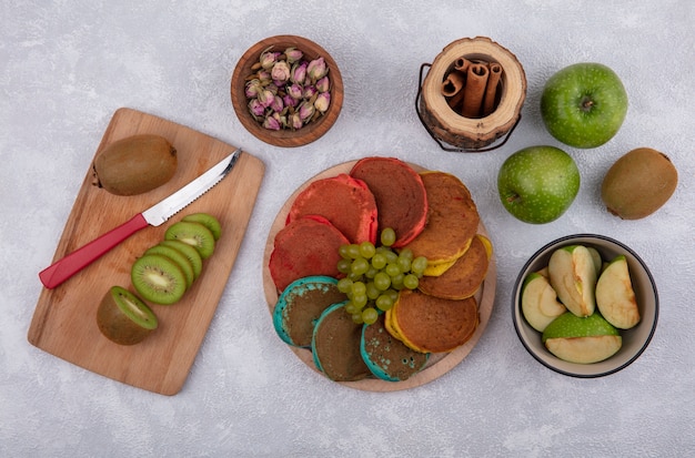 Top view colored pancakes with green grapes on a stand with green apples and kiwi with a knife on a board with cinnamon on a white background