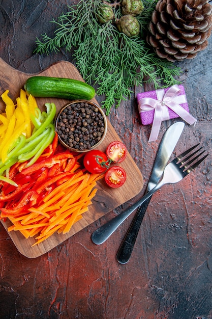 Top view colorful cut peppers black pepper tomatoes cucumber on cutting board knife and fork small gifton dark red table