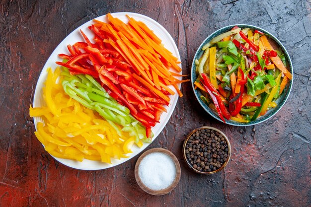 Top view colorful cut peppers on white plate vegetable salad in bowl black pepper salt garlic on dark red table