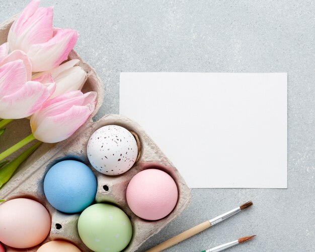 Top view of colorful easter eggs in carton with tulips and paper