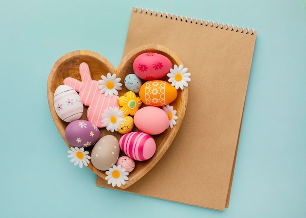 Top view of colorful easter eggs in heart-shaped plate with notebook