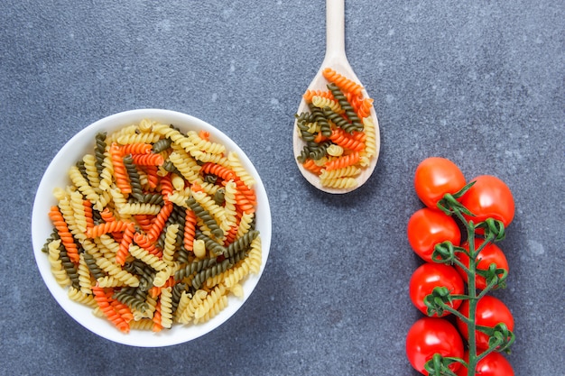 Free photo top view colorful macaroni pasta in a bowl and spoon with tomatoes on gray surface. horizontal