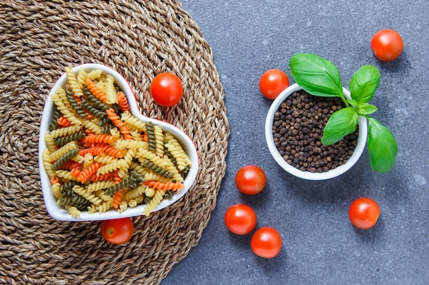 Top view colorful macaroni pasta in heart shaped bowl with tomatoes, black pepper on trivet and gray surface. horizontal