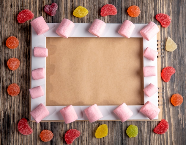 Free photo top view of colorful marshmallow arranged on an empty picture frame and marmalade candies on rustic