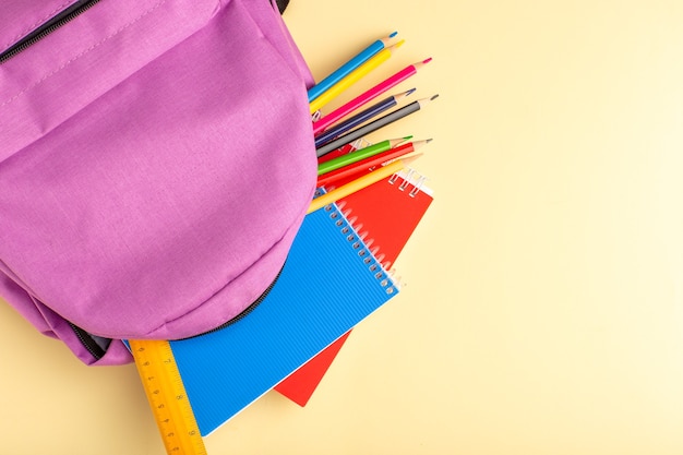 Top view colorful pencils with copybooks and purple bag on light-yellow wall school felt pen pencil book notepad