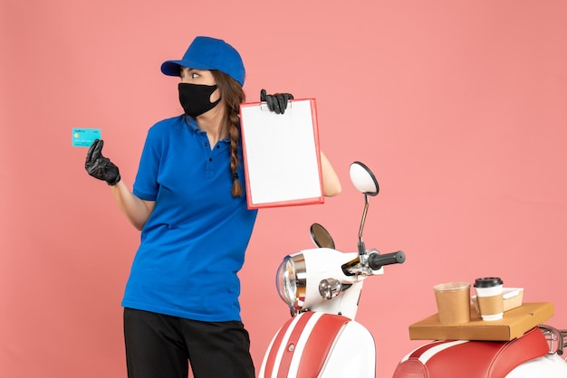 Free photo top view of concentrated courier girl wearing medical mask gloves standing next to motorcycle with coffee cake on it holding documents bank card on pastel peach color background