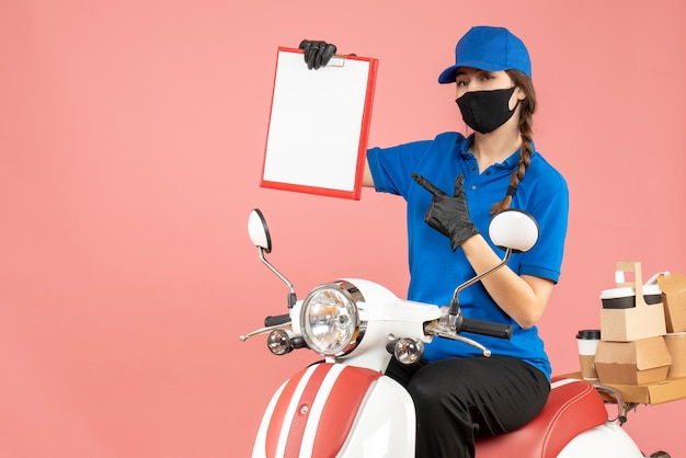 Free photo top view of confident courier girl wearing medical mask and gloves sitting on scooter holding empty paper sheet delivering orders on pastel peach background