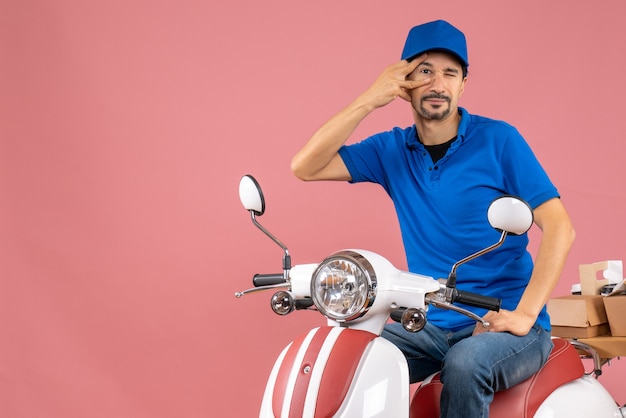 Top view of confident courier guy wearing hat sitting on scooter delivering orders on pastel peach background