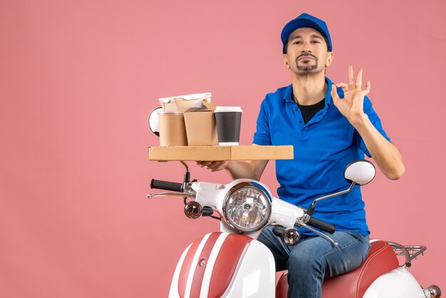 Top view of confident courier man wearing hat sitting on scooter making perfect gesture on pastel peach background