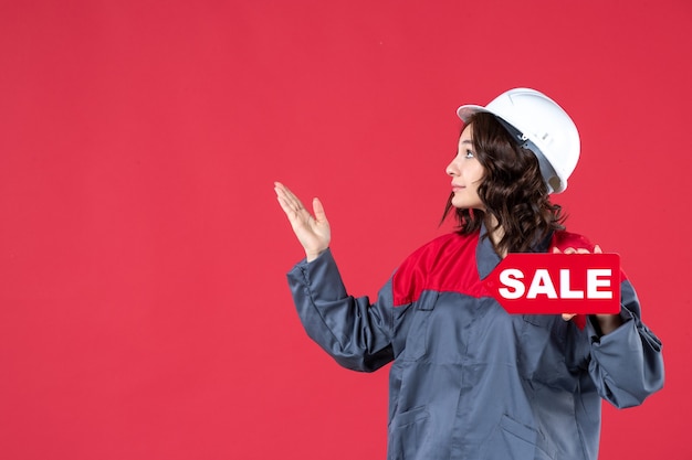 Free Photo top view of confident smiling female builder in uniform wearing hard hat and showing sale icon pointing up on the right side on isolated red background