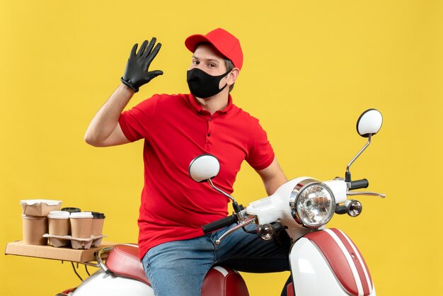 Top view of confident young adult wearing red blouse and hat gloves in medical mask delivering order sitting on scooter showing five on yellow background