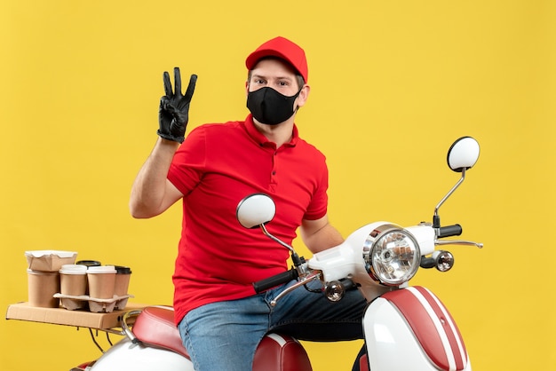 Top view of confident young adult wearing red blouse and hat gloves in medical mask delivering order sitting on scooter showing three on yellow background