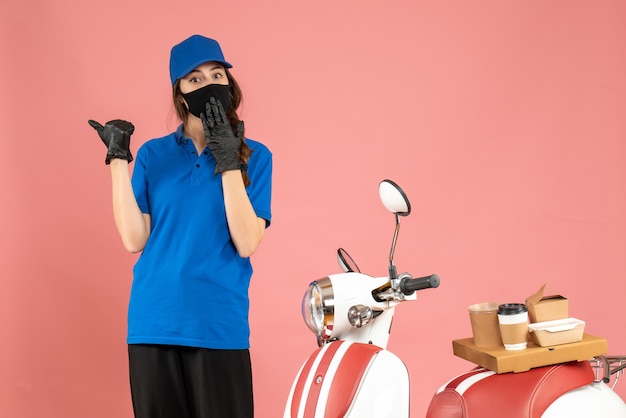 Top view of confused courier girl in medical mask standing next to motorcycle with coffee cake on it on pastel peach color background