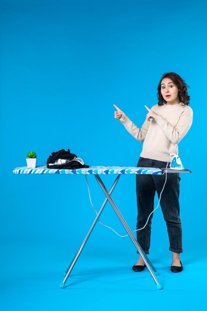 Top view of confused young girl standing behind the ironing board pointing something on blue wave background