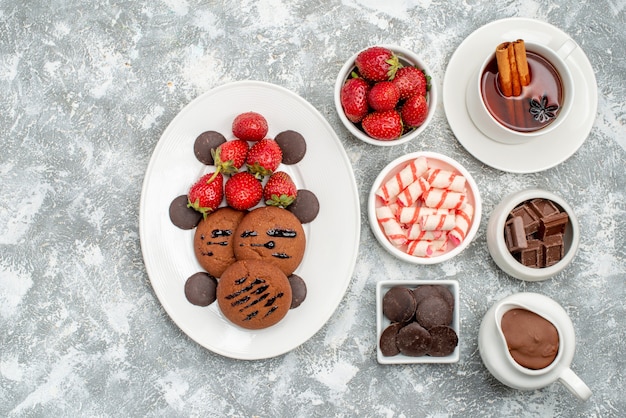 Top view cookies strawberries and chocolates bowls with cacao candies strawberries chocolates and tea with cinnamon at the right side of the grey-white table