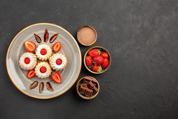 Free Photo top view cookies and strawberries cookies with strawberries on white plate next to bowls of strawberries chocolate and chocolate cream