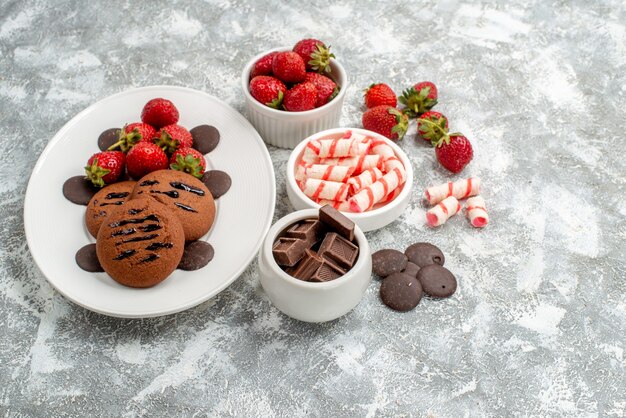 Top view cookies strawberries and round chocolates on the white oval plate bowls of candies strawberries chocolates on the grey-white table