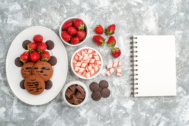 Top view cookies strawberries and round chocolates on the white oval plate bowls of candies strawberries chocolates and notebook on the grey-white table