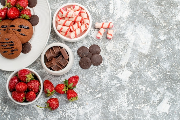 Top view cookies strawberries and round chocolates on the white oval plate bowls of candies strawberries chocolates at the top left of the grey-white table