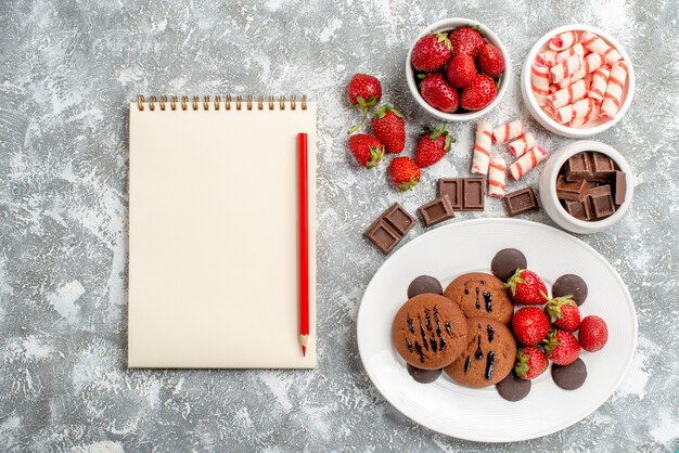 Top view cookies strawberries and round chocolates on the white plate and bowls of candies strawberries chocolates and notebook on the grey-white ground