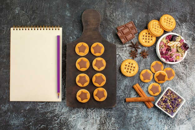 Top view of cookies on wooden platter with notebook and pen wit bowls of dry flowers on grey ground