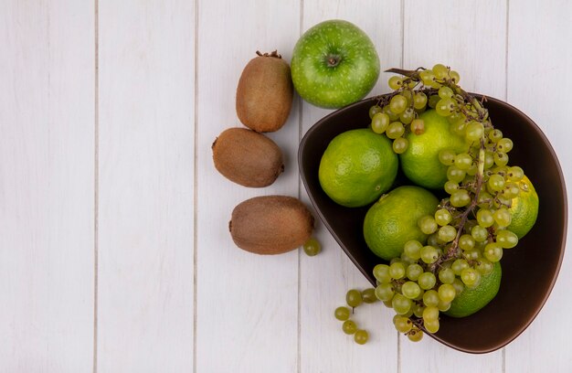 Top view copy space kiwi with apple and grapes with tangerines in a bowl on white wall