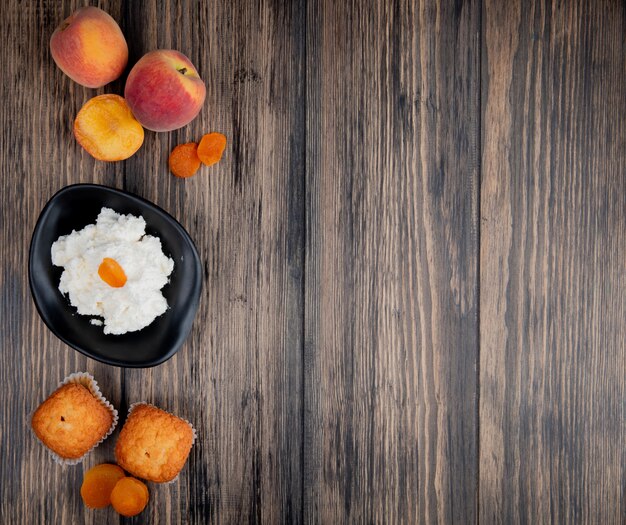 top view of cottage cheese in a black bowl with muffins fresh peaches and dried apricots on rustic wooden table with copy space