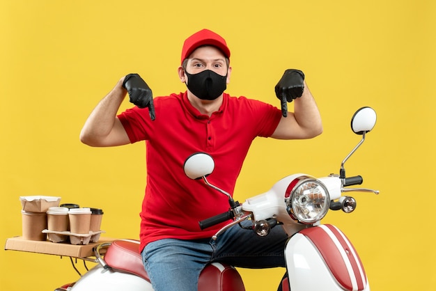 Top view of courier guy wearing red blouse and hat gloves in medical mask delivering order sitting on scooter pointing down