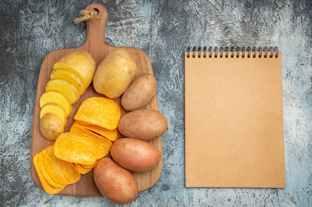 Free photo top view of crispy chips and uncooked potatoes on wooden cutting board and notebook on gray background