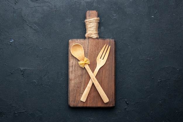 Top view crossed wooden spoon and fork on wooden serving board on dark table copy place