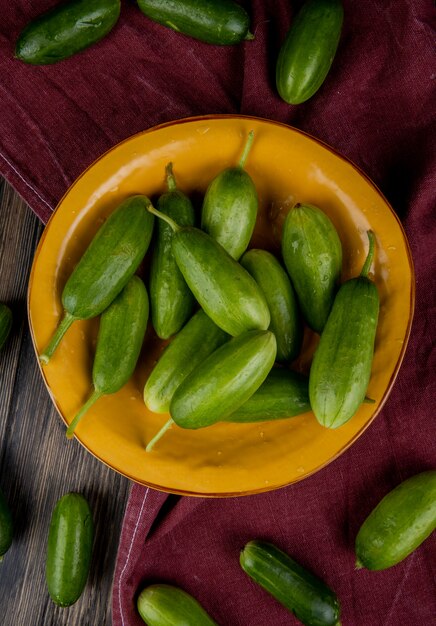 Top view of cucumbers in plate with other ones on bordo cloth and wooden surface