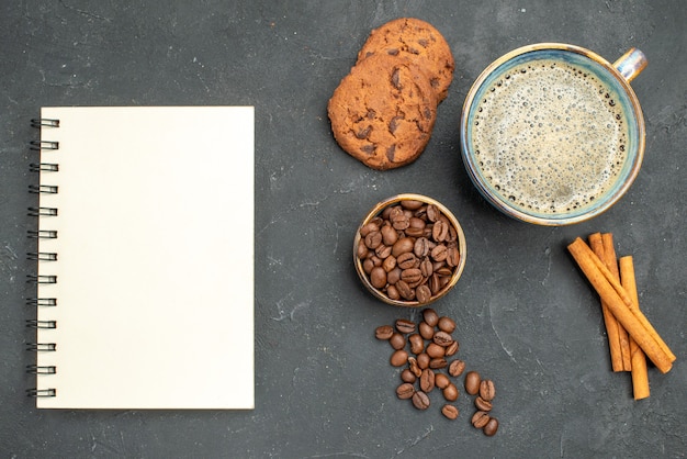 Top view a cup of coffee bowl with coffee seeds cinnamon sticks biscuits a notebok on dark isolated background