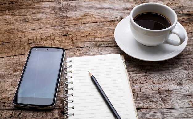 Top view of a cup of coffee, smart phone and black pencil on empty notebook over wooden table.