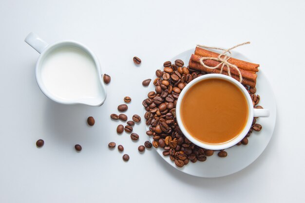 Top view a cup of coffee with coffee beans and dry cinnamon on saucer and with milk, on white surface