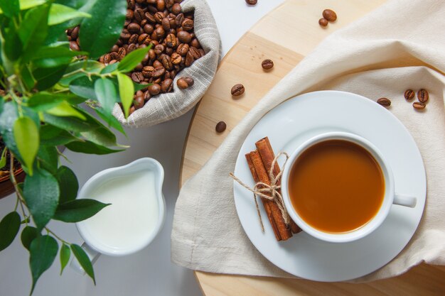 Top view a cup of coffee with coffee beans in a sack, plant, milk, dry cinnamon on platform and white surface vertical