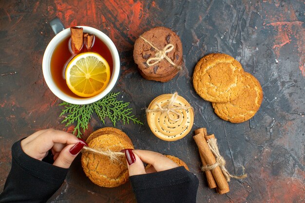 Top view cup of tea cinnamon sticks different cookies tied with rope in female hand on dark red table