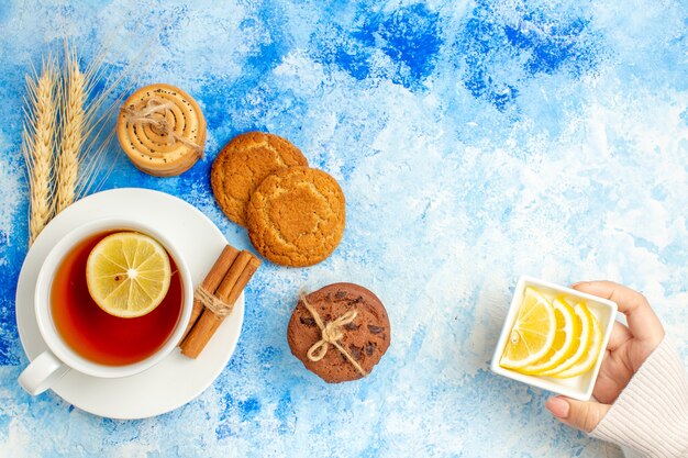 Top view cup of tea cookies lemon slices in bowl in woman hand on blue table free space