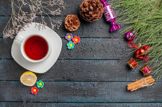 Top view a cup of tea a cup of black tea on the white saucer next to the lemon cinnamon sticks spruce branches with Christmas toys and cones on the table