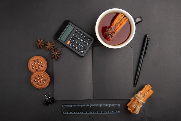 Top view cup of tea flavored by cinnamon and anise calculator notepad biscuits pen ruler on dark table