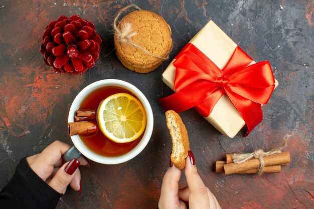 Top view cup of tea flavored by lemon and cinnamon and cookie in female hand xmas gift red pinecone cinnamon sticks cookies on dark red table