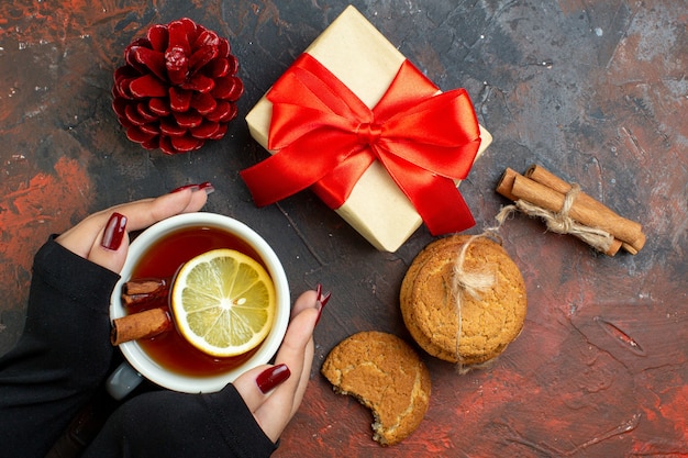 Free photo top view cup of tea flavored by lemon and cinnamon in female hand xmas gift red pinecone cinnamon sticks cookies on dark red table