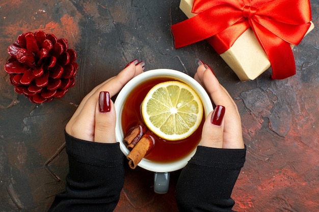 Top view cup of tea flavored by lemon and cinnamon in female hand xmas gift red pinecone on dark red table