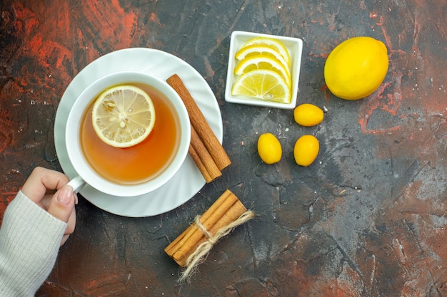 Free photo top view cup of tea flavored by lemon in female hand cumcuat lemon slices in small bowl cinnamons on dark red table